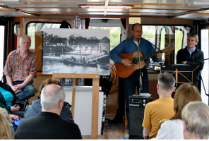 Musiker spielt Gitarre vor Publikum auf einem Boot, Schwarzweißfoto im Hintergrund
