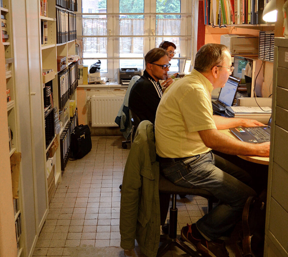 Drei Personen arbeiten an Computern in einem schmalen Archivraum.