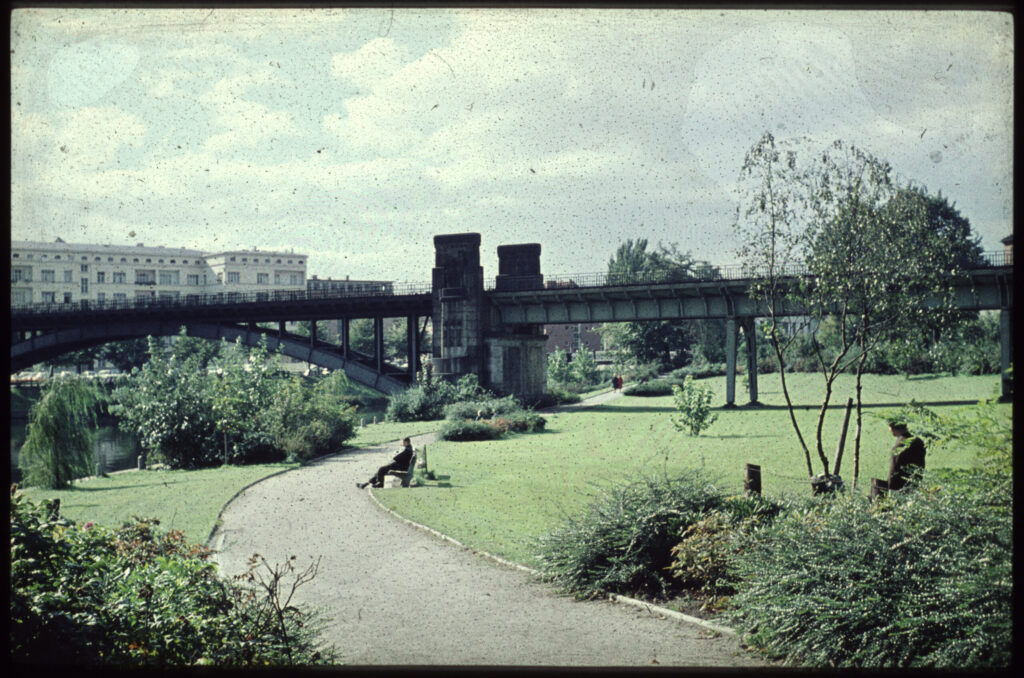 Grünanlage mit Wegen, Büschen und Brücke über Kanal, Menschen auf Bänken